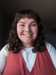 A selfie of Katherine smiling. She has wavy brown shoulder-length hair, and she's wearing a pink dress with a white long-sleeve shirt underneath.